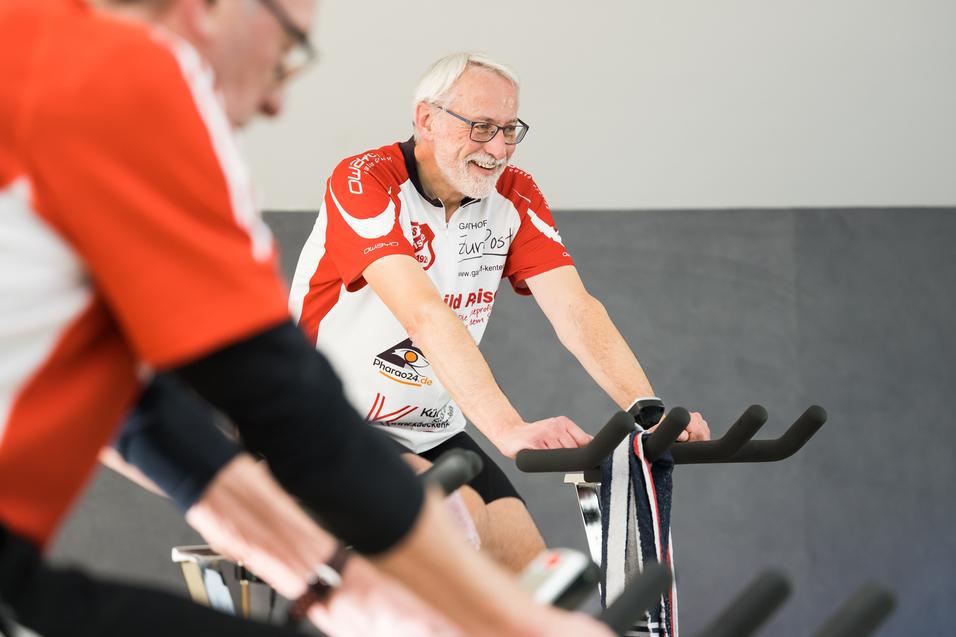 Zwei Radfahrer in Trainingskleidung lächeln auf stationären Fahrrädern während eines Fitnesskurses in einem Studio.