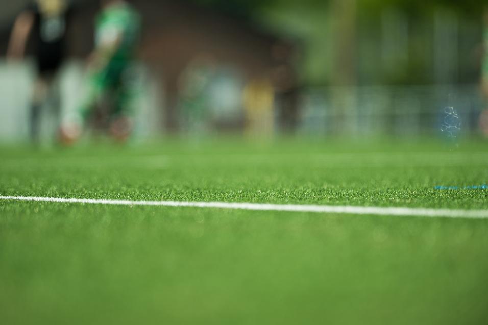 Nahaufnahme von grünem Kunstrasen mit einer weißen Linie; unscharfe Silhouetten von Spielern im Hintergrund.