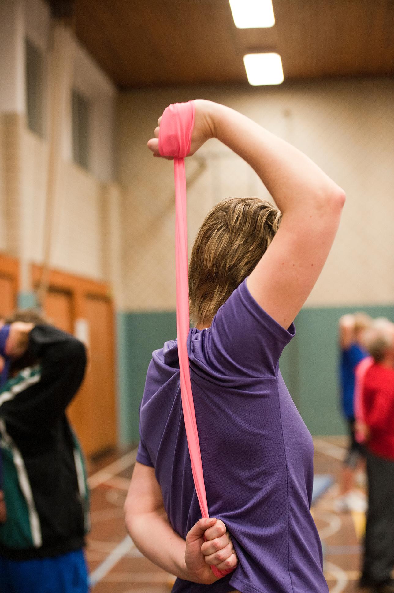 Frau in lila T-Shirt dehnt mit einem pinken Widerstandsband ihren Arm in einer Sporthalle. Hintergrund mit anderen Personen.