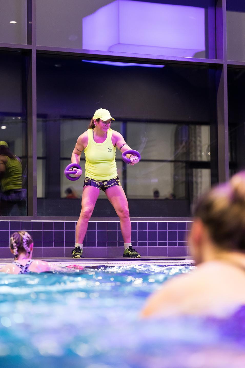 Frau in neonfarbener Sportbekleidung mit Hanteln steht am Rand eines Schwimmbeckens, im Hintergrund schwimmende Teilnehmerinnen.